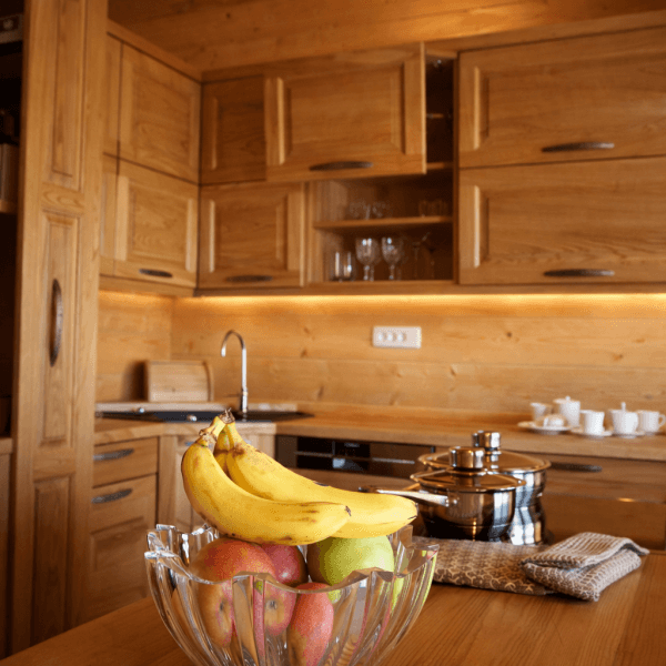 Staklena posuda s voćem, uključujući banane i jabuke, na drvenom stolu u kuhinji.